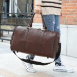 Un uomo in strada che trasporta una borsa da viaggio in finta pelle marrone per le sue cinghie. Sotto di essa pende una tracolla marrone. L'uomo è in piedi su un marciapiede della strada, dietro di lui c'è la facciata di una casa con una finestra.