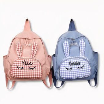 2 zainetti personalizzati con i nomi di Ylla e Kathlee. Uno è blu, l'altro rosa e hanno un disegno di orecchie di coniglio sul davanti, su uno sfondo grigio chiaro